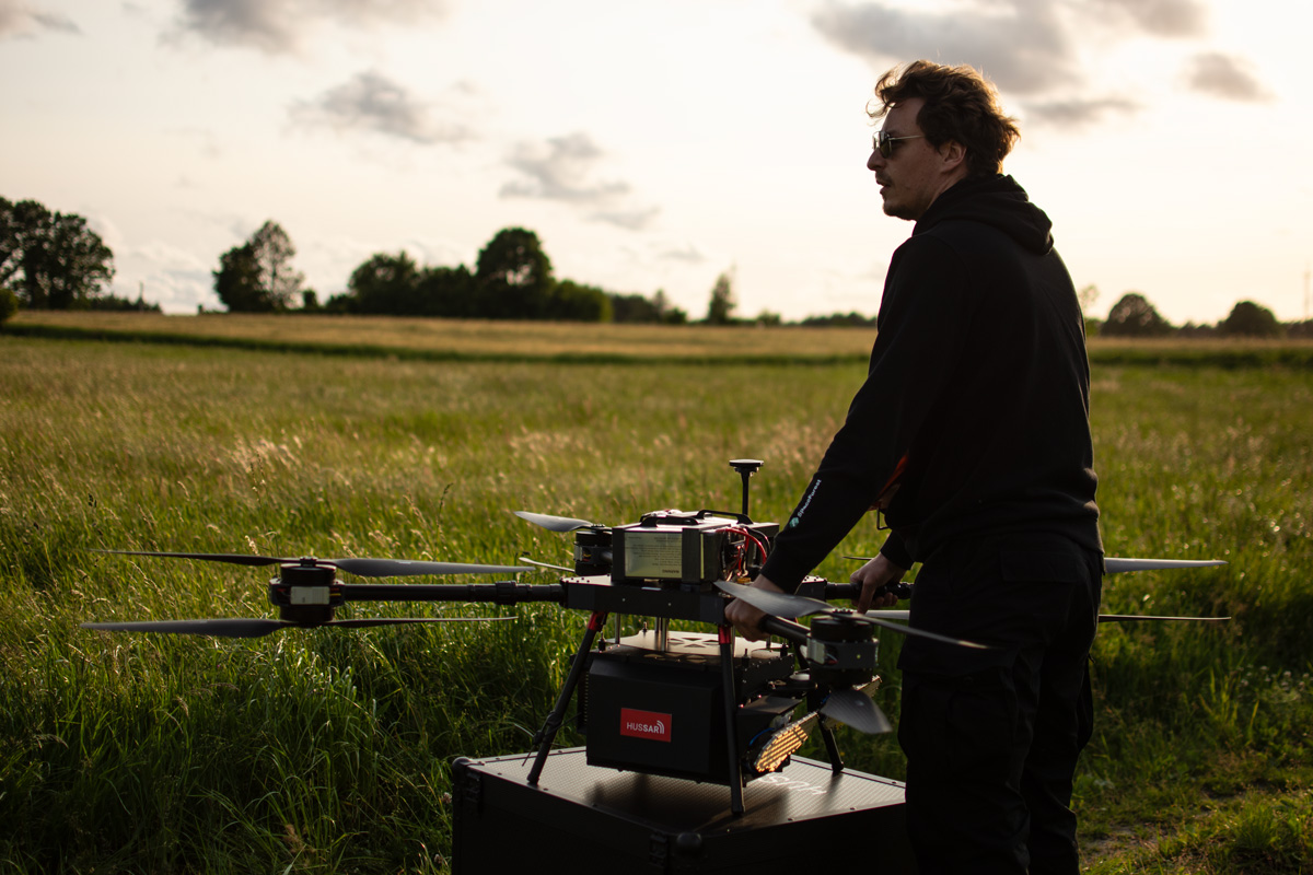 An operator overseeing the HUSSAR SAR drone standing on its transport case, set against a sunset backdrop in a field.
